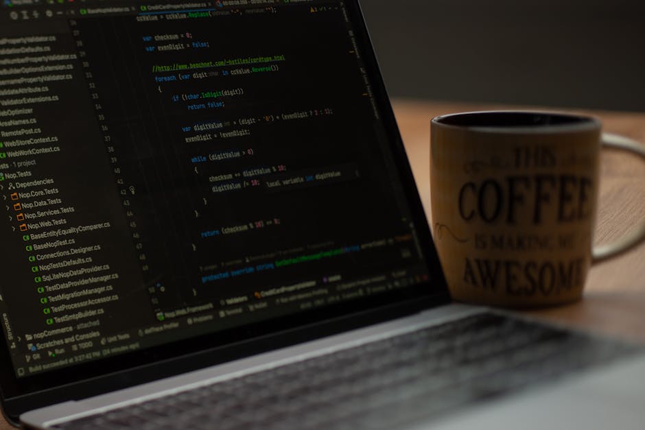 A close-up of a laptop screen displaying lines of computer code with colorful syntax highlighting, positioned on a wooden desk. Next to the laptop, there is a ceramic coffee mug with the text 'This co
