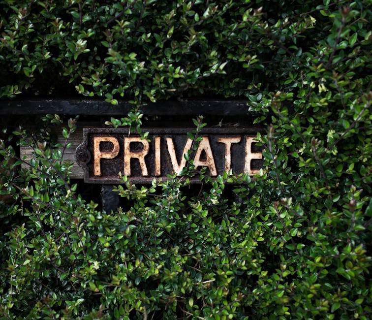 A close-up view of a weathered, rust-colored sign with the word 'PRIVATE' embossed in capital letters, mounted on a dark wooden board. The sign is partially obscured by dense, glossy green foliage con