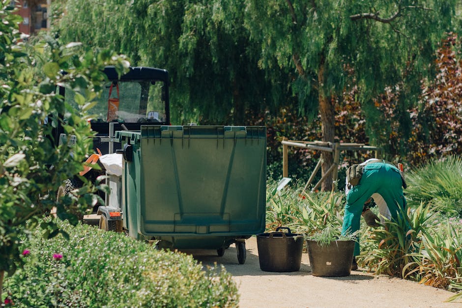 A gardener wearing teal work clothing is crouched down in a lush, well-maintained garden bed, carefully tending to a variety of green plants and foliage. Next to the gardener, two black buckets are pl