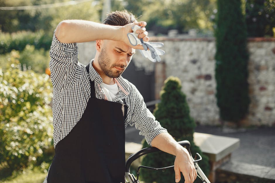 A gardener wearing teal work clothing is crouched down in a lush, well-maintained garden bed, carefully tending to a variety of green plants and foliage. Next to the gardener, two black buckets are pl